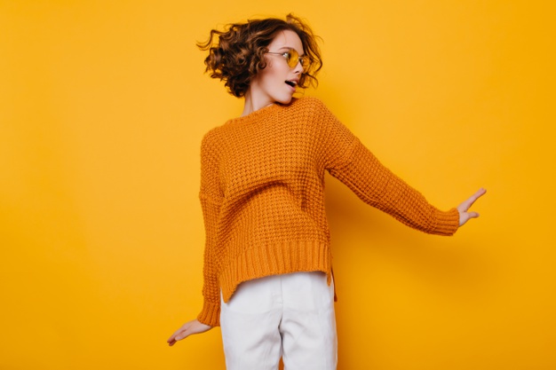 mujer con jersey mostaza y pantalón blanco sobre fondo amarillo.